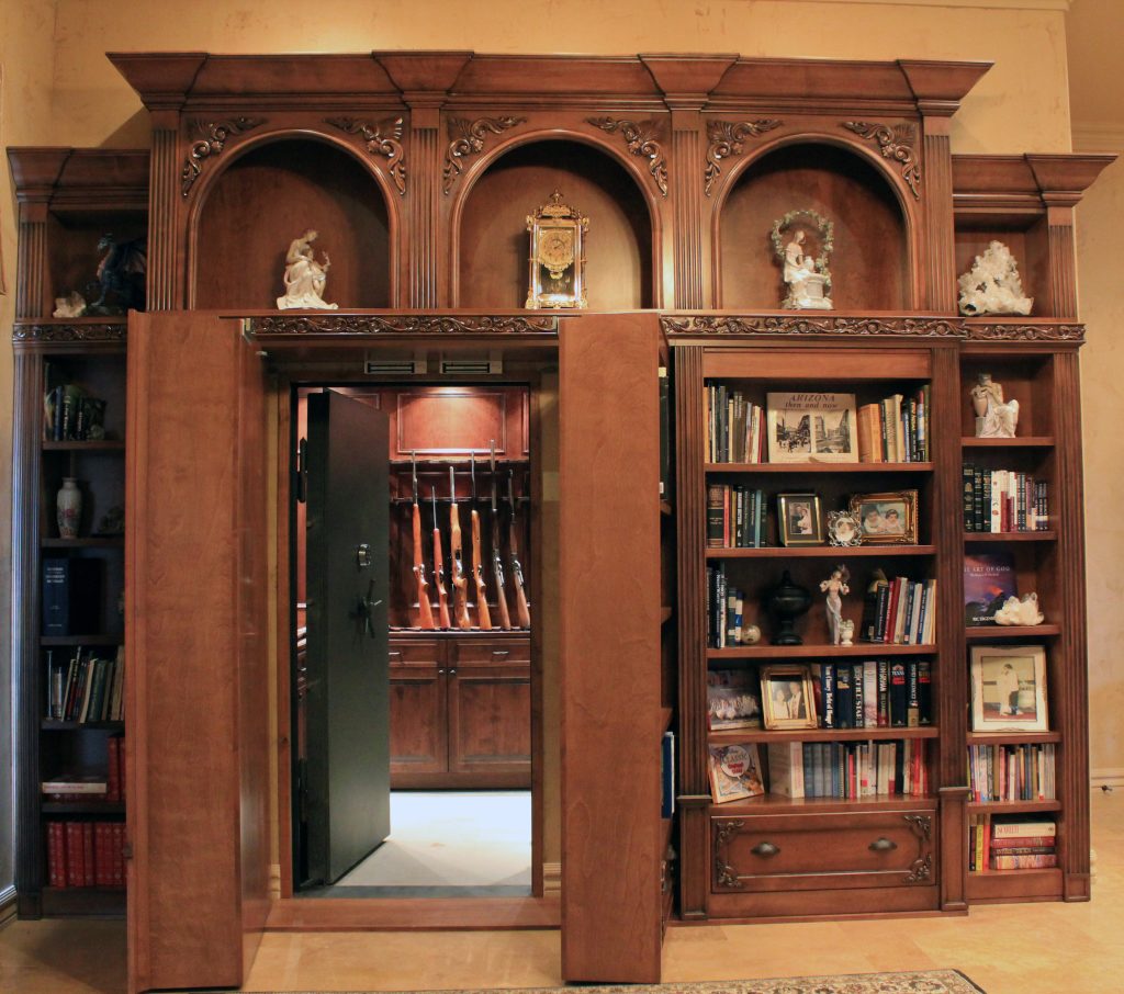 An ornate wooden bookcase with decorative archways and bookshelves opens to reveal a hidden vault door leading to a gun storage room with rifles displayed inside