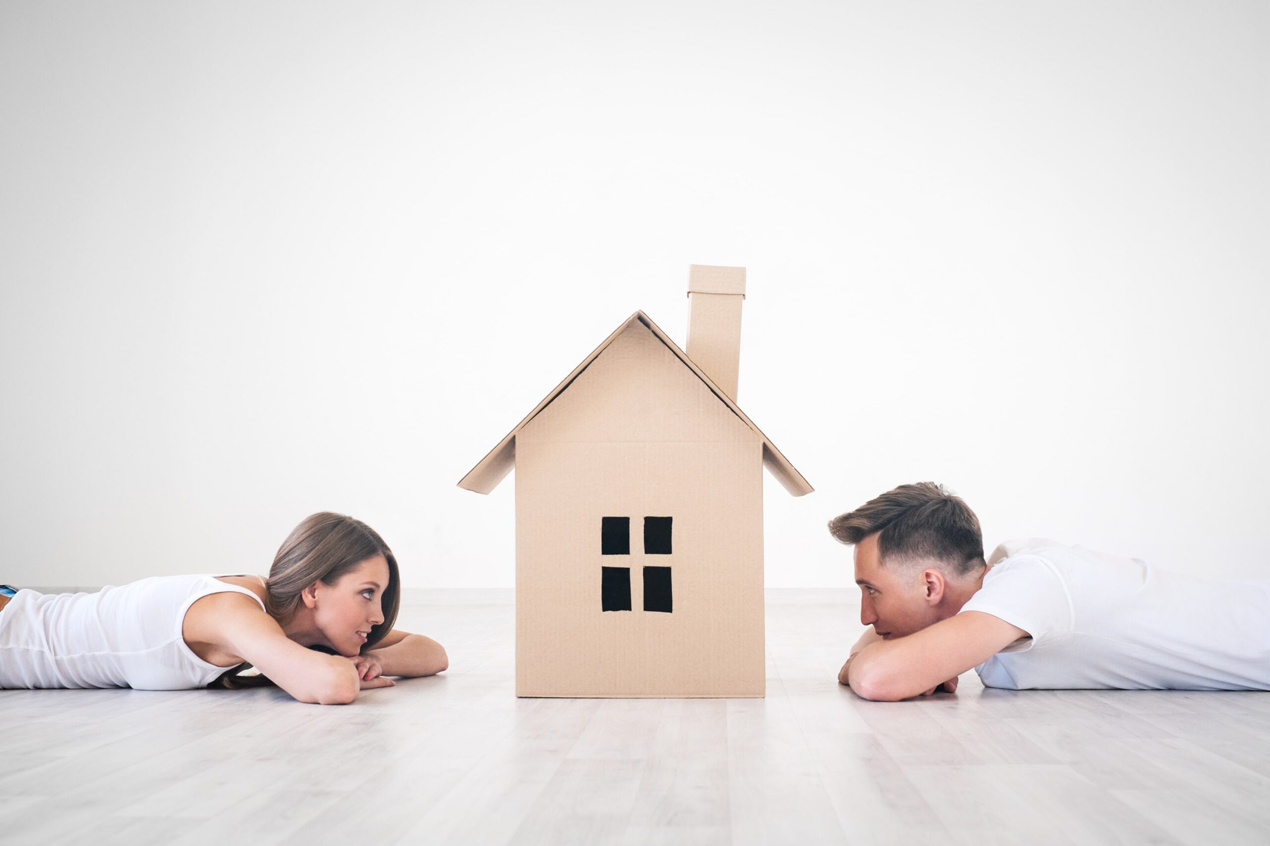 two people lying on the floor looking at a cardboard house.