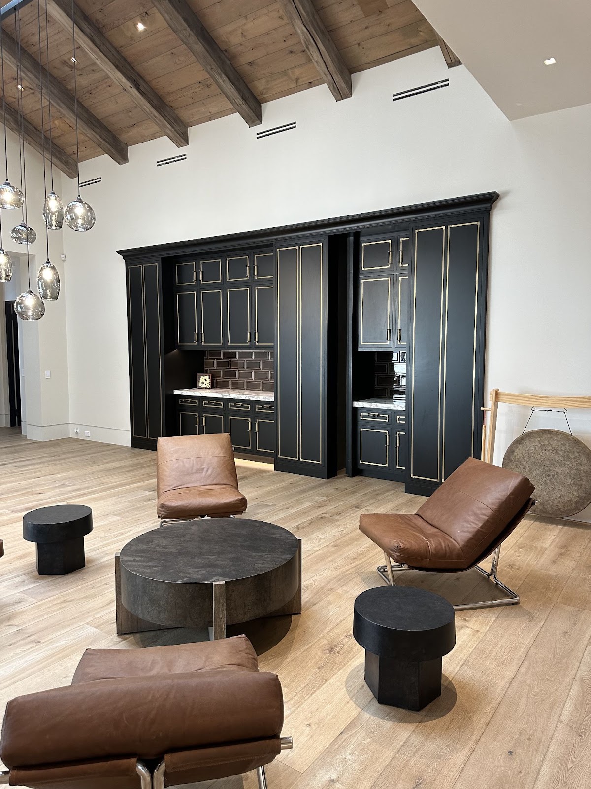 Modern living space with exposed wood beam ceiling and leather lounge chairs, featuring a hidden door integrated into a black and gold-trimmed cabinetry wall that opens to reveal a concealed room.