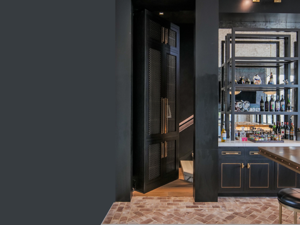 Elegant hidden door built into a tall black cabinet with gold hardware, partially open to reveal a secret staircase behind a home bar setup with shelves and bottles.