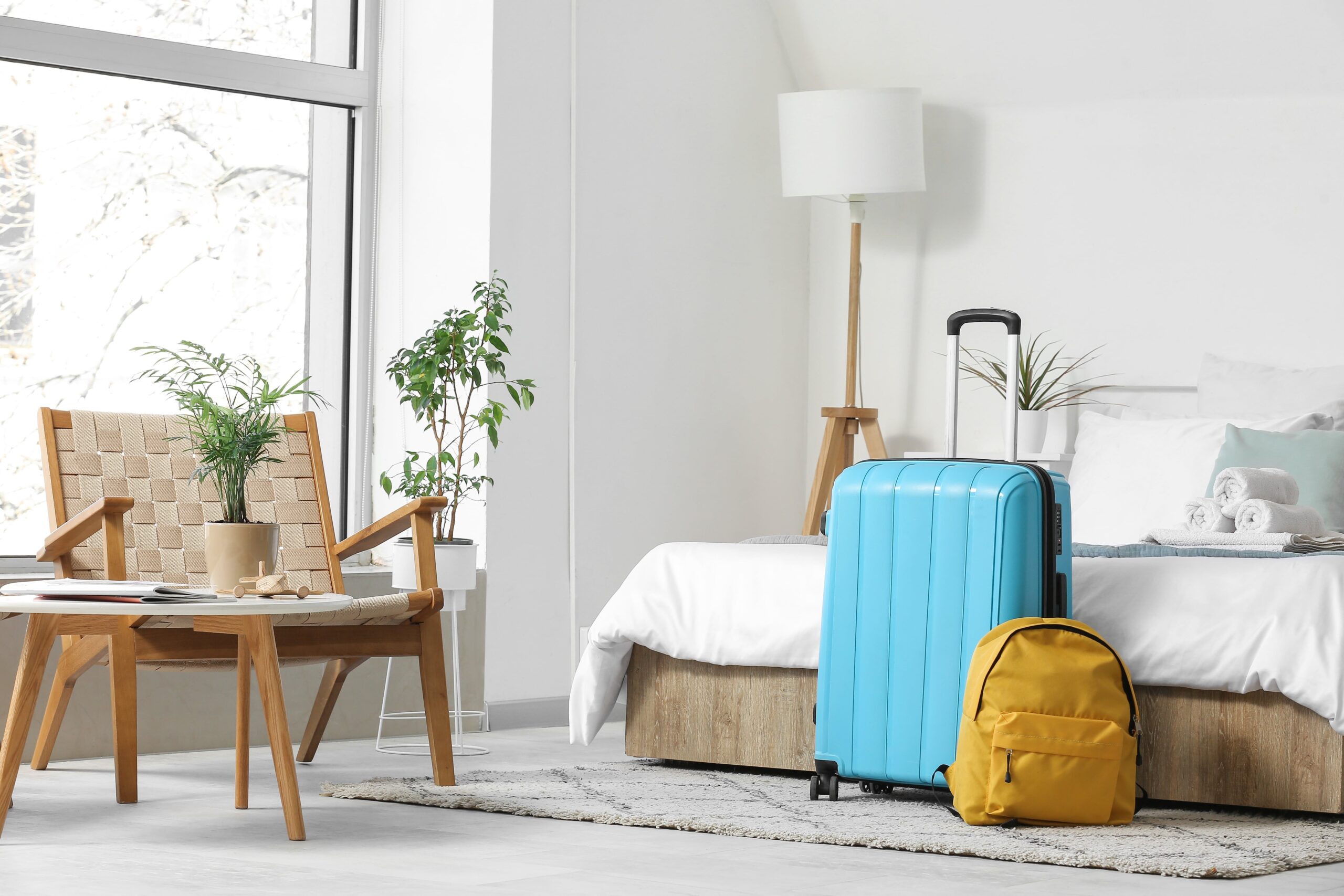 Modern bright guest bedroom with large windows, a blue suitcase, yellow backpack, and light wood furniture.