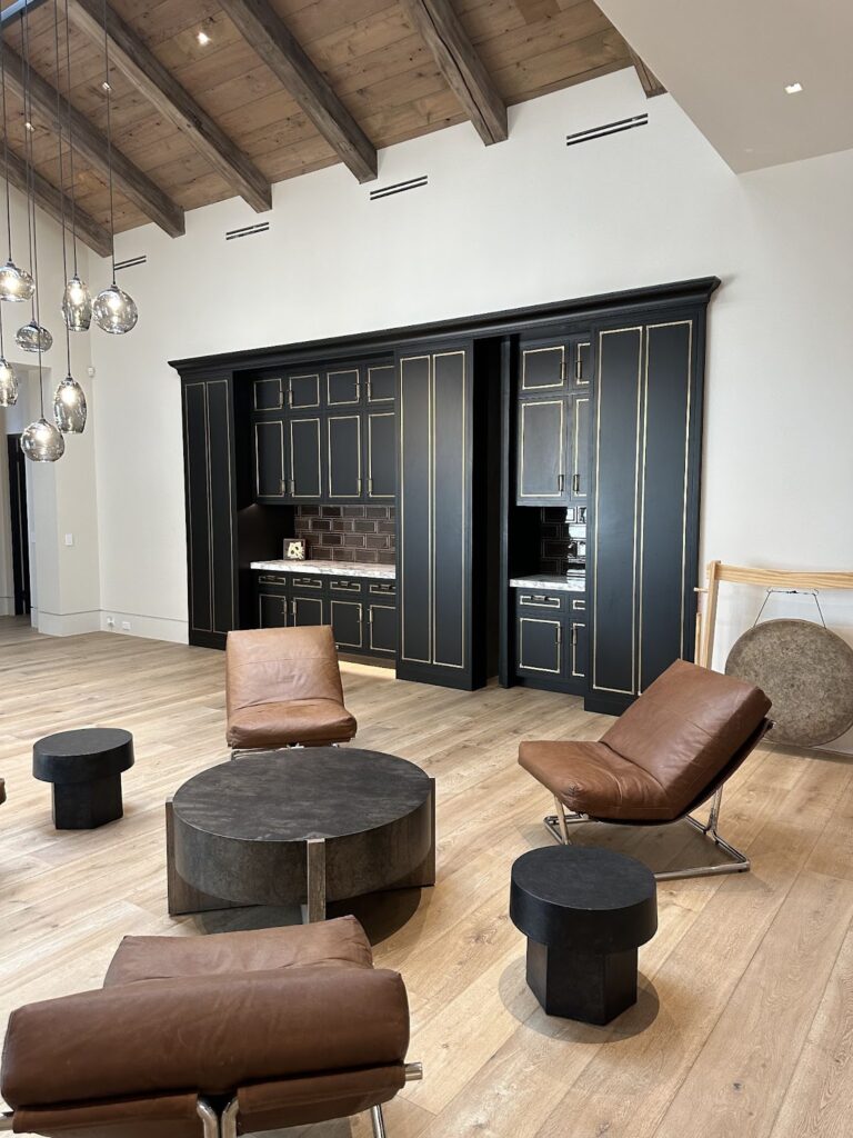  Modern living room with vaulted wood ceiling, leather lounge chairs, and hidden kitchen cabinetry featuring black panels with gold trim, blending into the wall. Hidden door designed by Creative Home Engineering.
