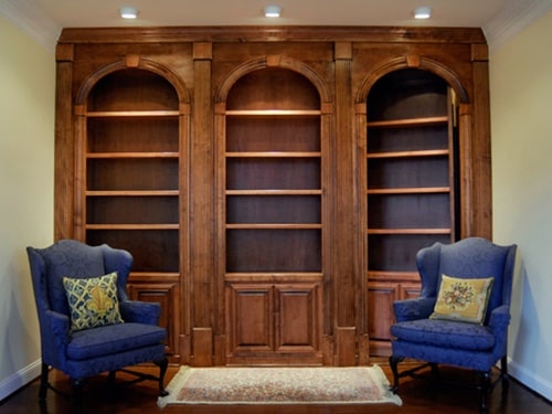 Traditional wood-paneled bookcase with three arched sections, flanked by two blue armchairs with decorative pillows, and a rug on the hardwood floor. Security bookcase hidden door designed by Creative Home Engineering
