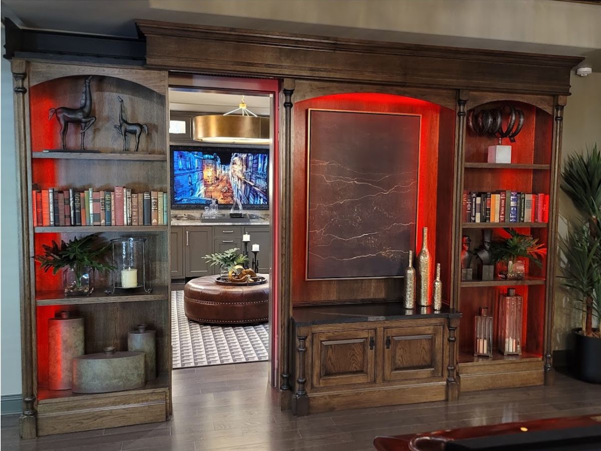 Custom sliding bookcase secret door with red backlighting, sliding open to reveal a modern entertainment room with TV, ottoman, and bar cabinetry.