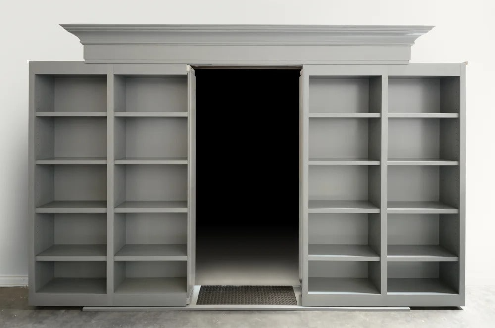 Gray double sliding bookcase secret door with crown molding, open in the center to reveal a hidden passage. 