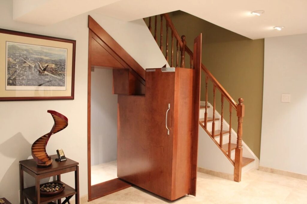 A custom hidden door built seamlessly into wood paneling beneath a staircase, blending into the surrounding trim.
