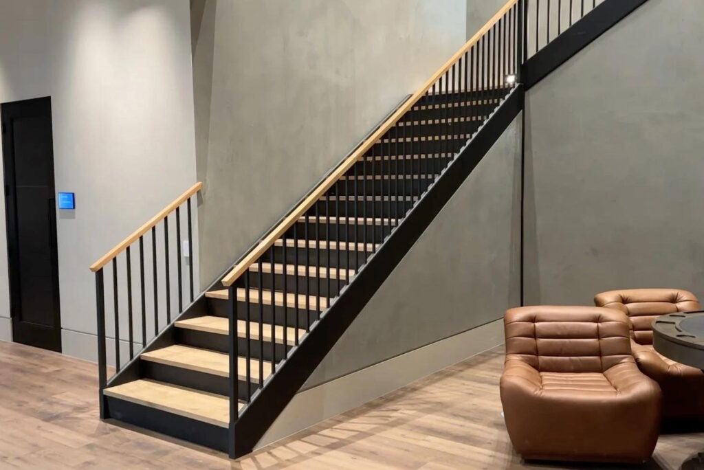 Closed lifting staircase with black metal frame and wood treads, concealing a hidden space beneath in a modern interior with neutral walls, wood flooring, and leather seating nearby.