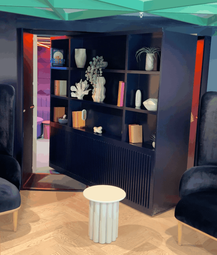 A dark bookshelf filled with books and decor swings open as a hidden door, revealing a colorful room behind. Two black chairs and a small white table sit in front on a wooden floor.