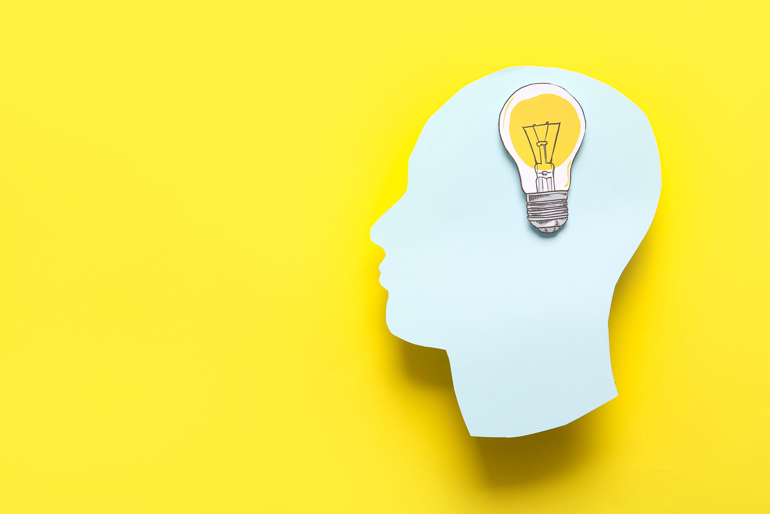 A blue paper cutout of a human head is shown in profile on a yellow background, with a drawing of a light bulb placed where the brain would be, symbolizing an idea or creativity.